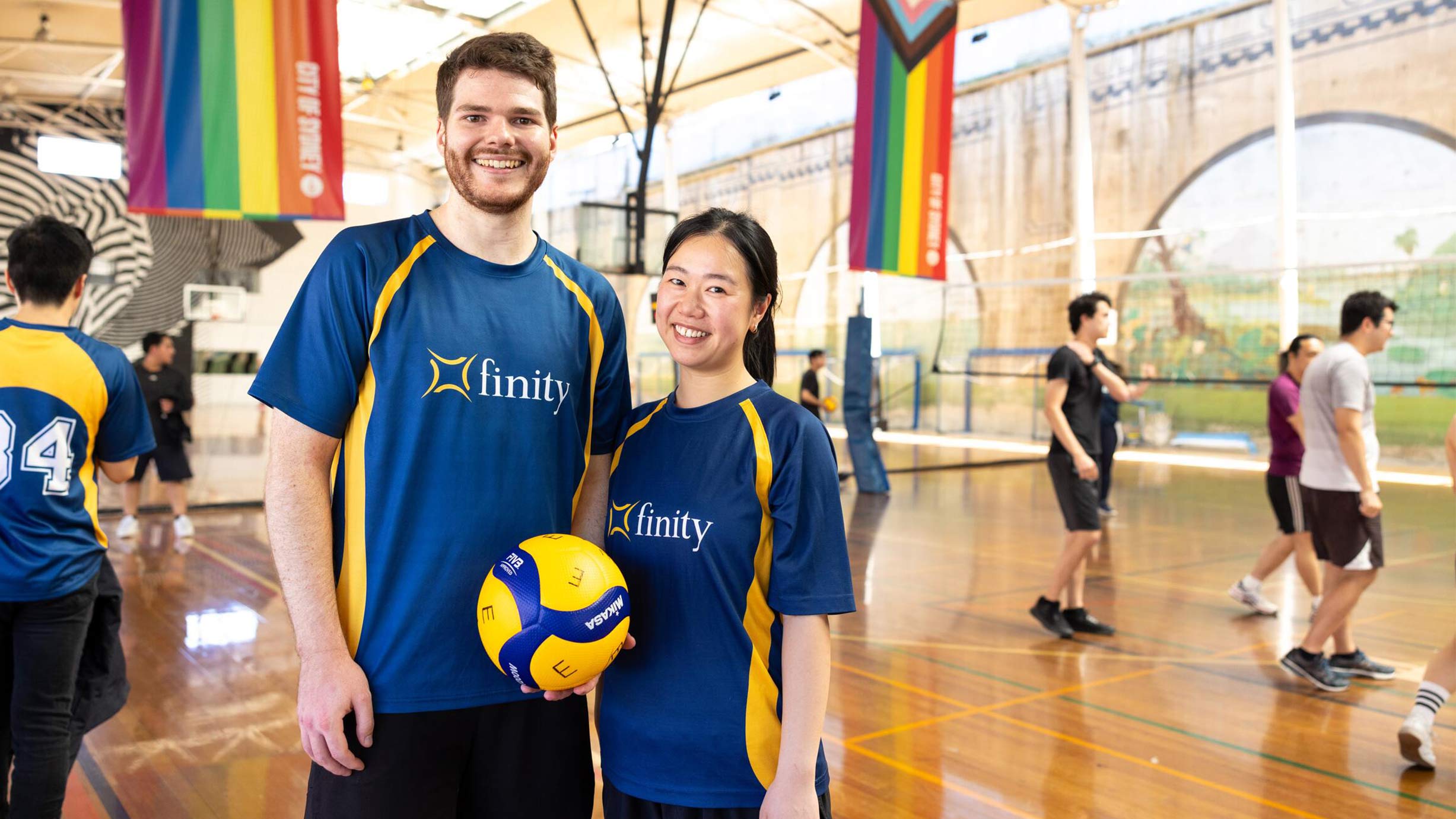 Two people in matching blue and yellow sports jerseys hold a volleyball and smile at an indoor gym with other players in the background.